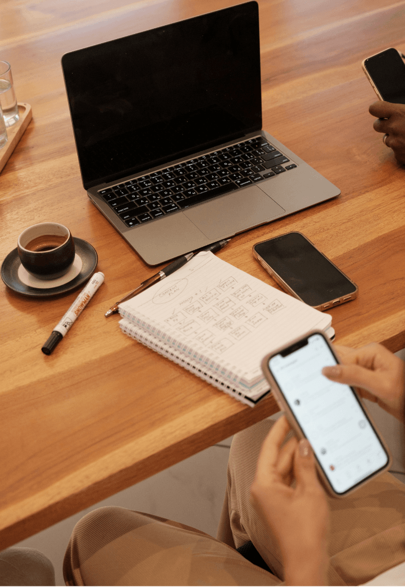 Une personne sur son téléphone assis devant son bureau, une tasse de café, un ordinateur et son carnet de notes.