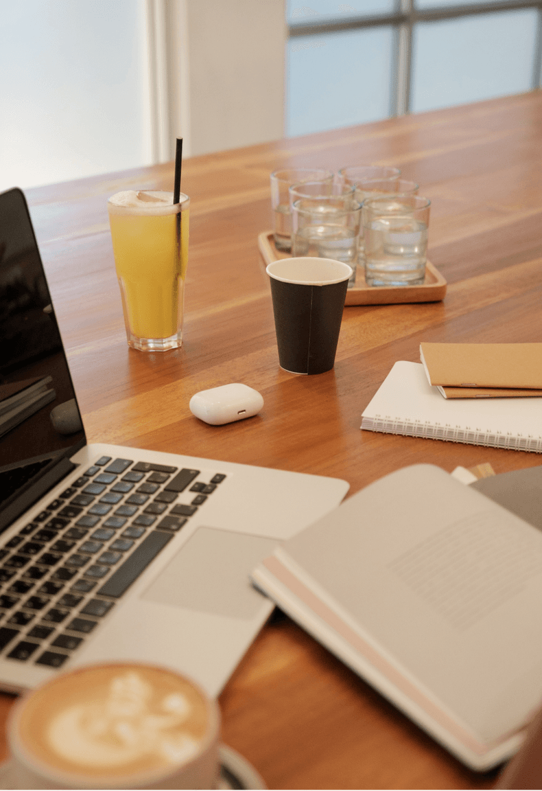 Un bureau avec une boite de crayons, un ordinateur, un jus de fruit, un café, un cahier de notes.